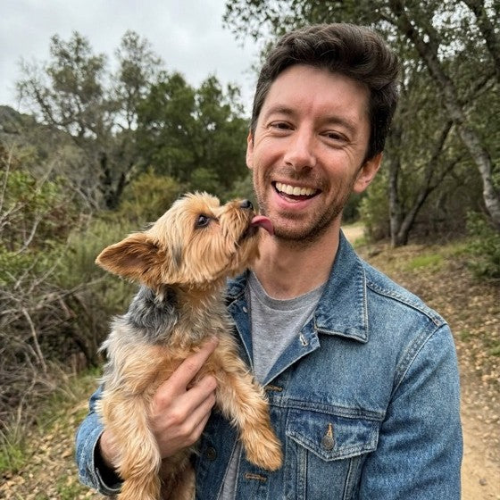 Gus, who runs Lorvo, smiling outdoors while a small dog licks his cheek